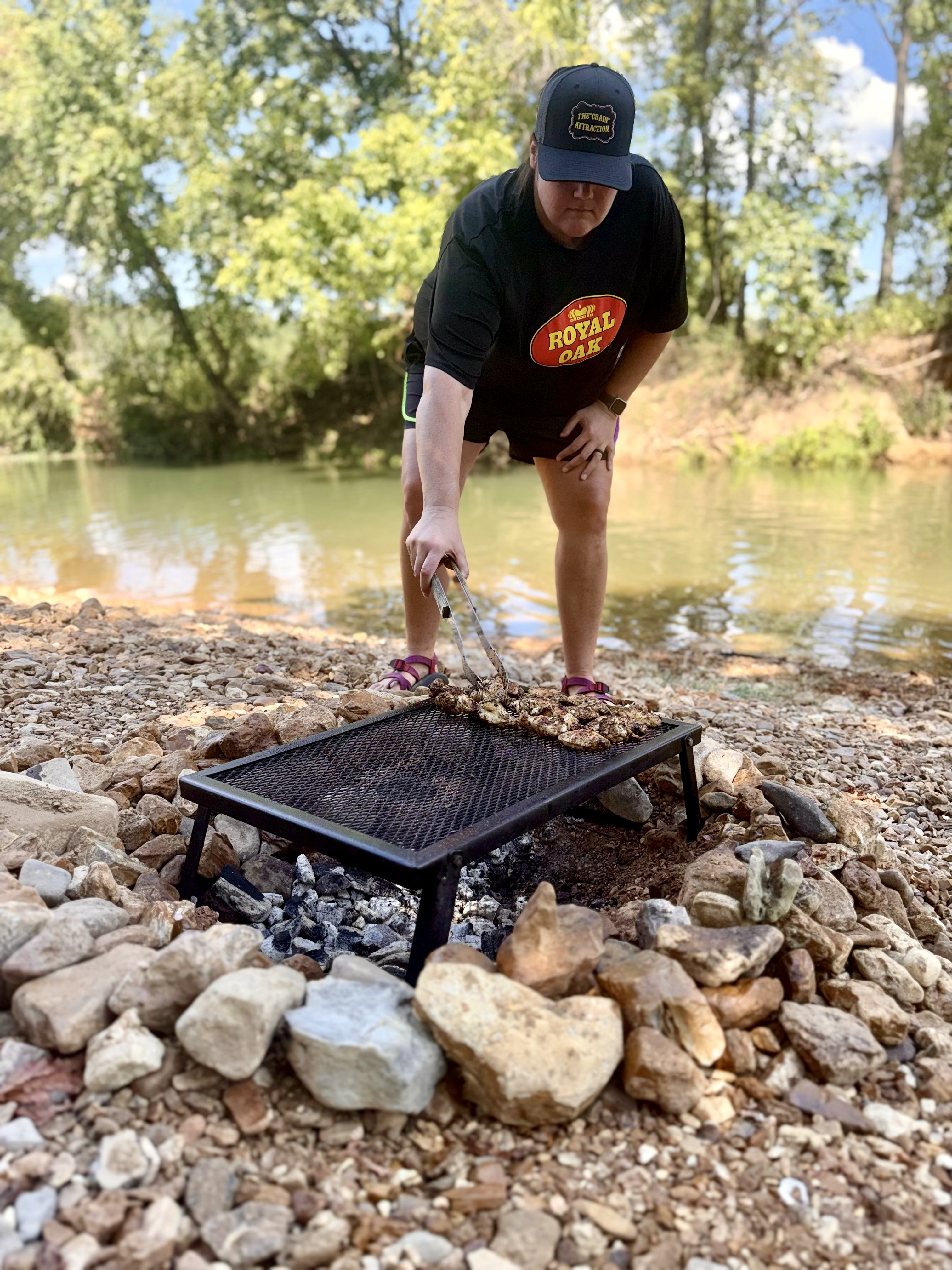 The Crain Attraction Grilling chicken wings over open Royal Oak charcoal with The CRAIN Attraction seasoning on an outdoor riverbank. Caption: Keep it simple: clean heat, fresh air, and coals dialed in. Great barbecue doesn't require an over-complicated setup